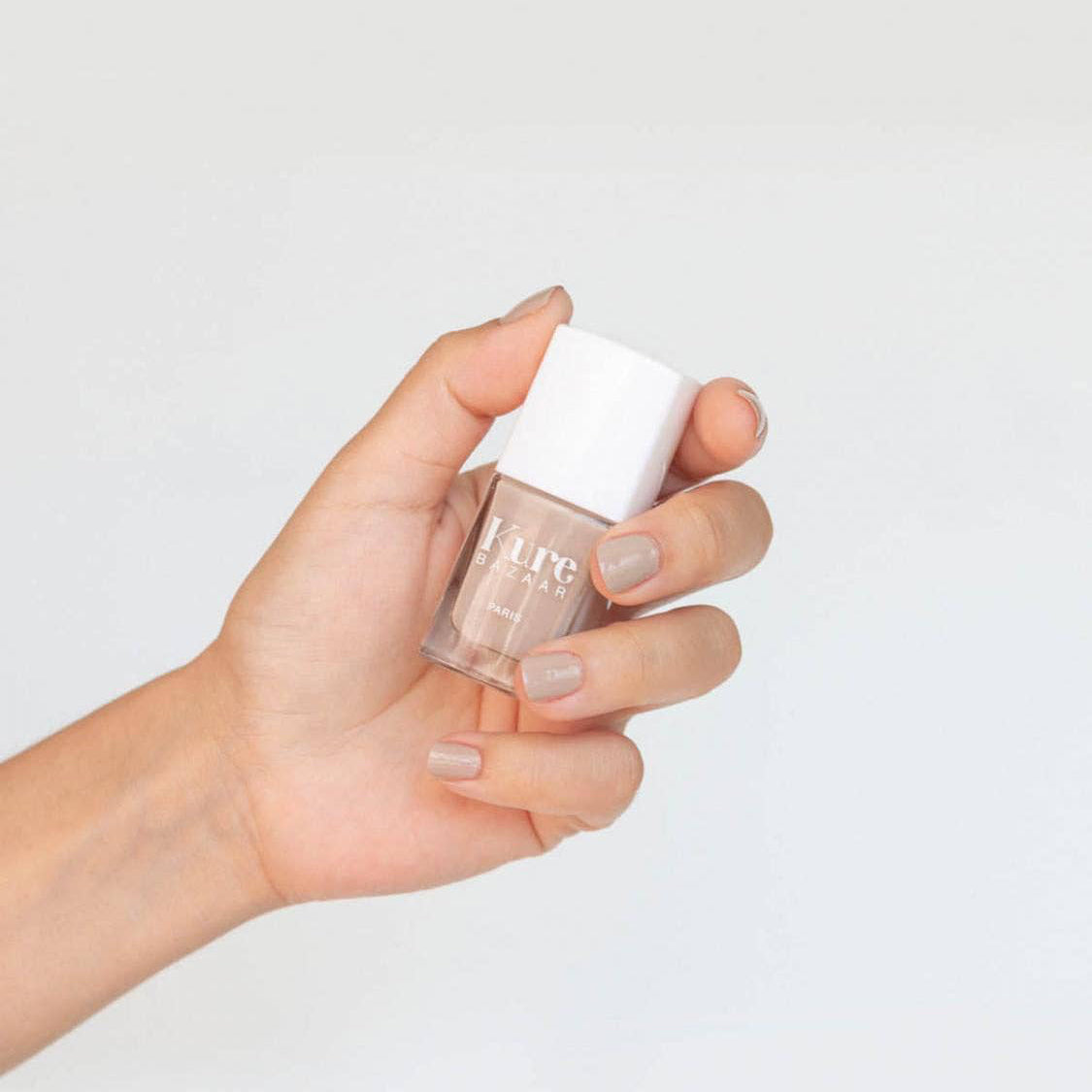 Hand holding a bottle of nail polish with a white cap on a light gray background