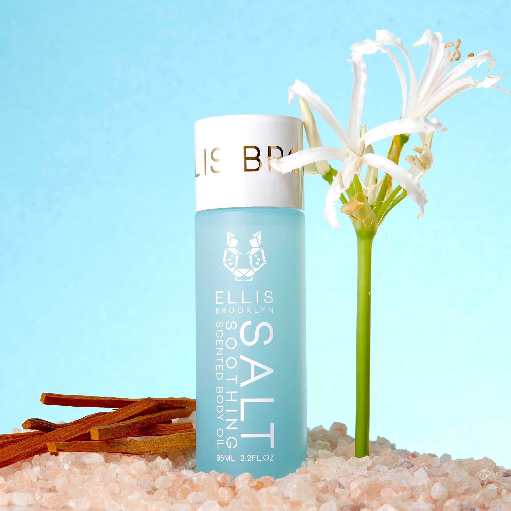 Ellis Brooklyn Scented Body Oil bottle with flowers and salt on a light blue background