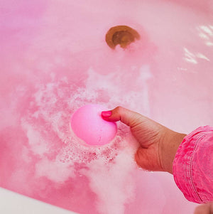 Hand holding a pink bath bomb over a bathtub filled with pink water.