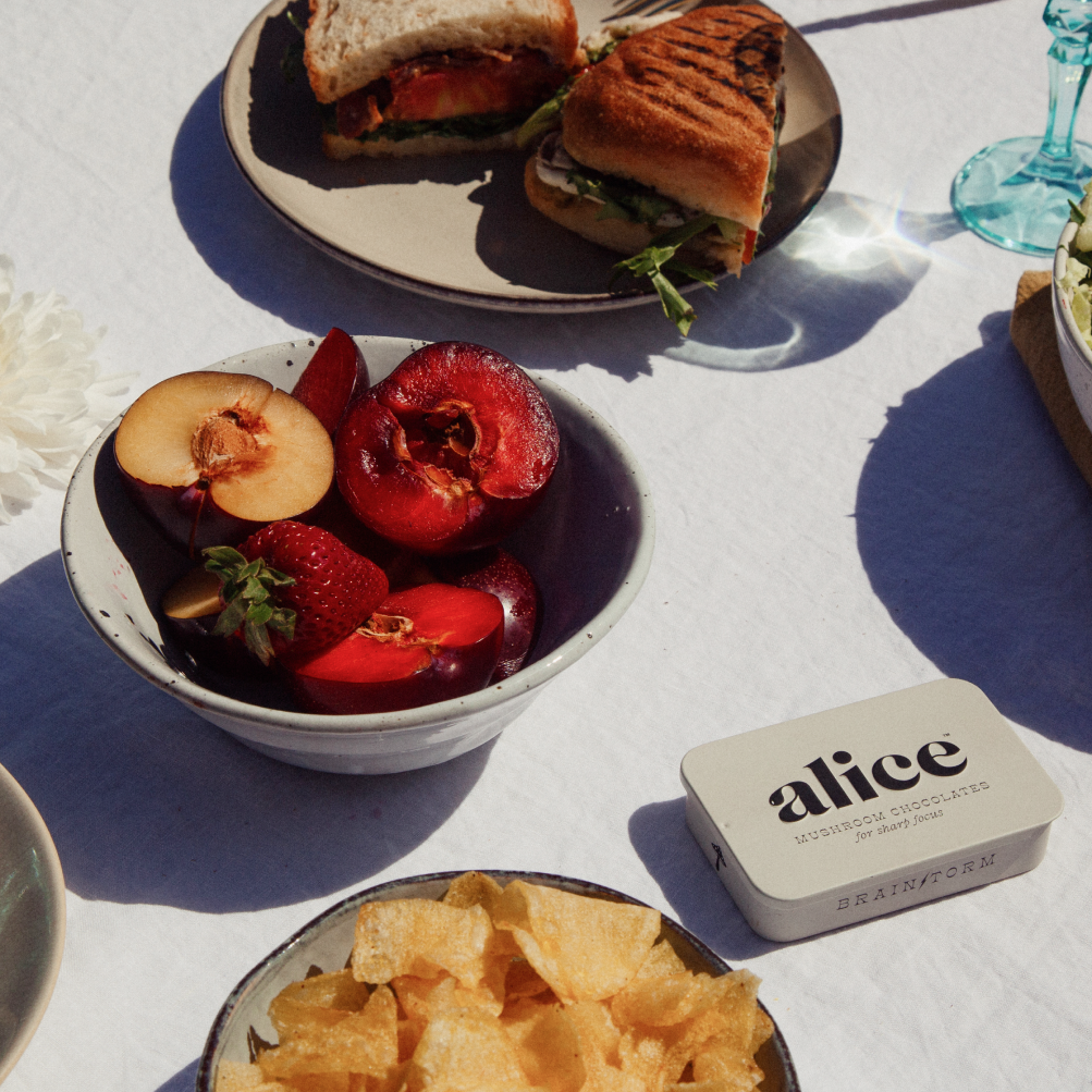 Outdoor setting with a bowl of fruit, chips, and a sandwich on a white tablecloth.