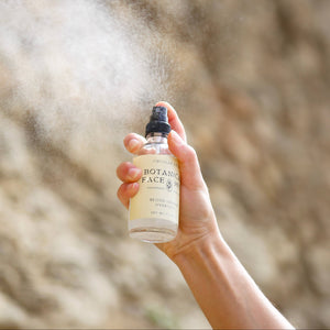 Hand holding a spray bottle labeled 'Botanical Face Wash' against a natural background