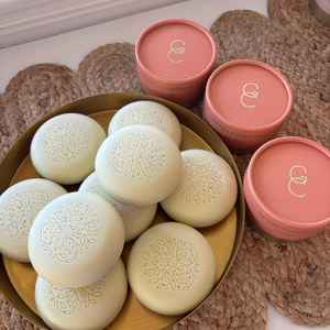 White round lotion bars with gold embossed design in a gold bowl, surrounded by pink containers with a logo.