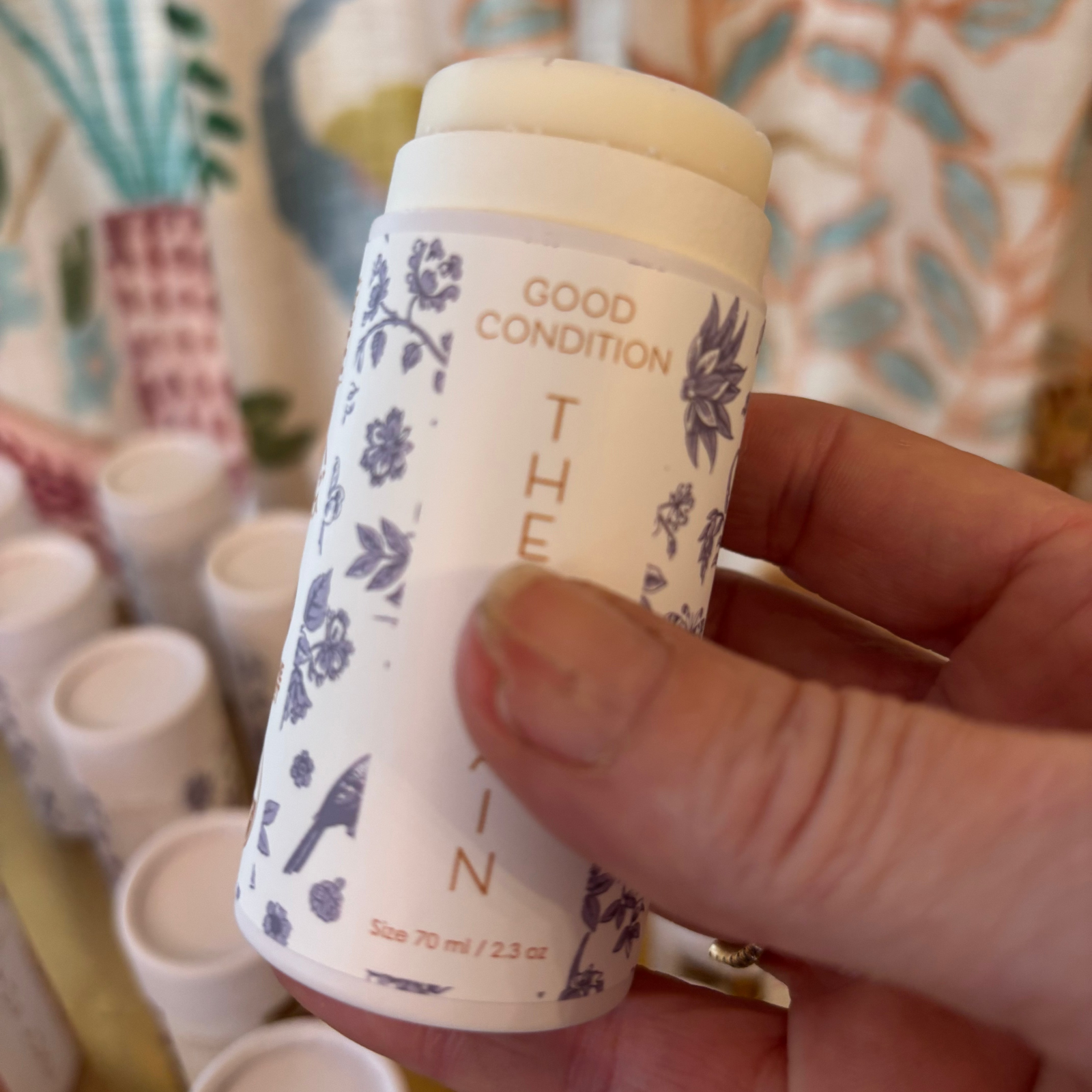 Hand holding a cylindrical product labeled 'The Plain' with floral design against a colorful background.