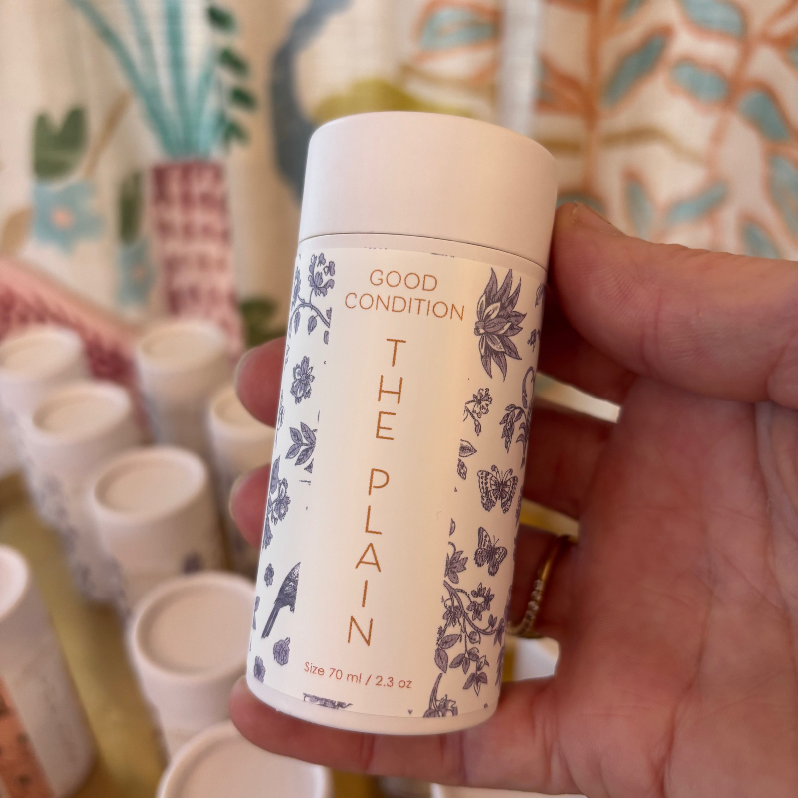 Hand holding a cylindrical product labeled 'The Plain' with floral design against a colorful background.