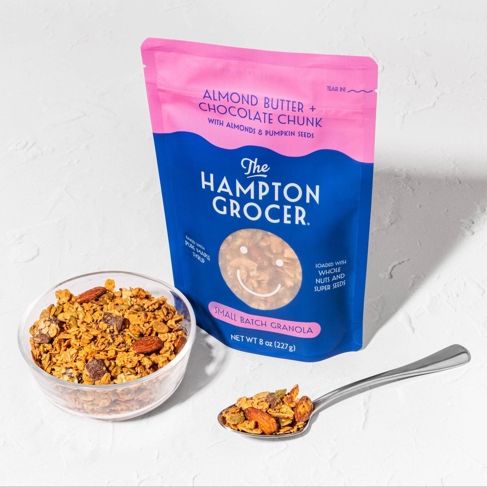 The Hampton Grocer almond butter chocolate chex mix with a bowl and spoon on a white background