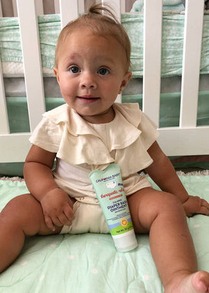 Baby sitting on a crib with a diaper rash cream bottle