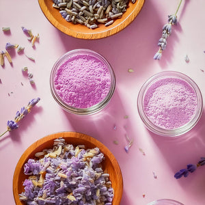 Two glass jars of pink powder with wooden bowls of lavender flowers and buds on a pink background.
