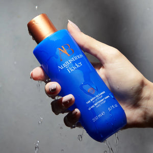 Hand holding a blue bottle of Augustinus Bader body cleanser with water droplets around it.