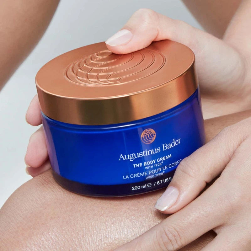 Blue jar of Augustinus Bader body cream held by a hand with a neutral background
