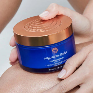 Blue jar of Augustinus Bader body cream held by a hand with a neutral background