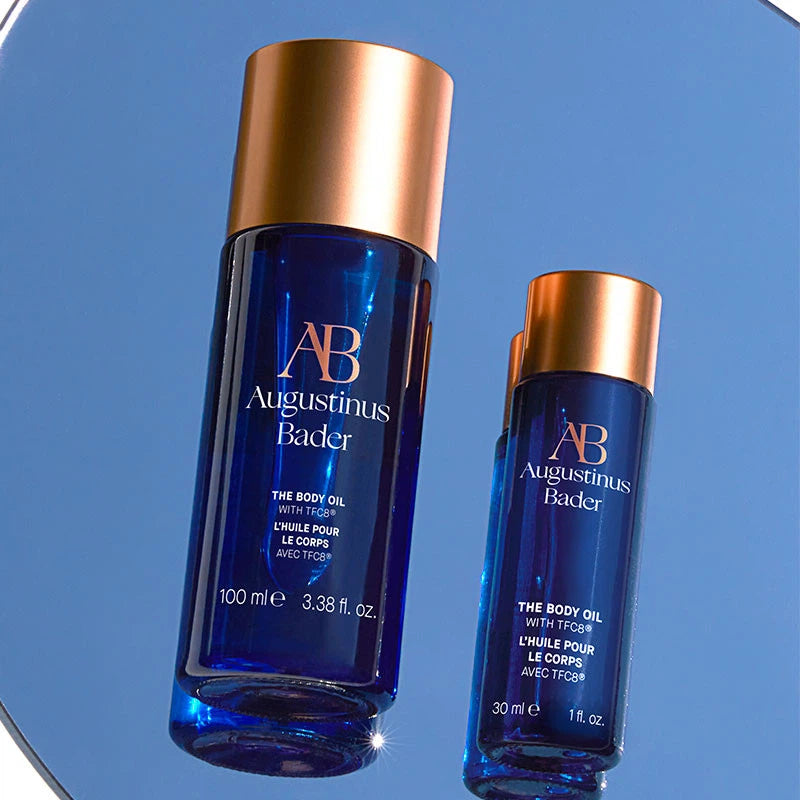 Two bottles of Augustinus Bader body oil on a reflective blue surface