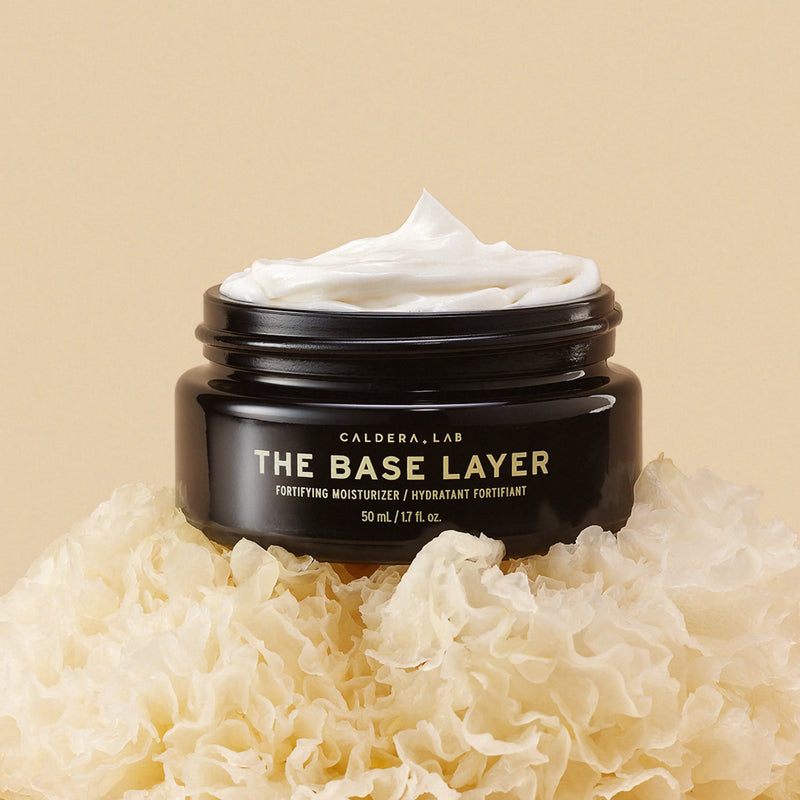 A black jar with a gold and white label reading 'THE BASE LAYER' by Caldera + Lab, indicating a nourishing day moisturizer.