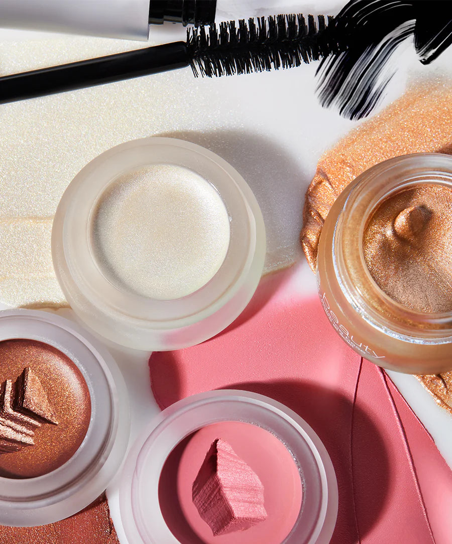 Makeup products including a mascara, jars of cream, and a compact on a pink surface.