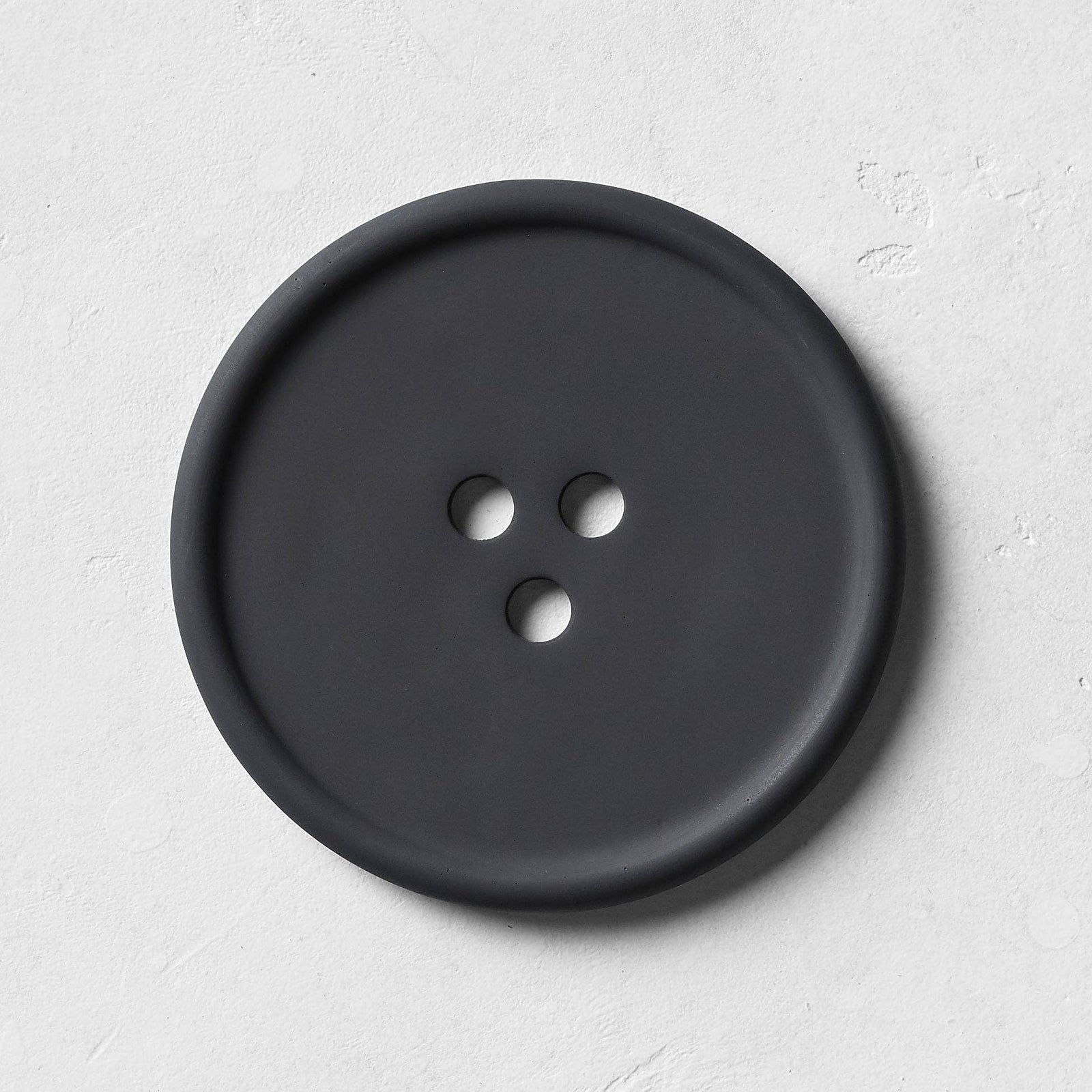 A round black concrete tray with a button-like shape, featuring subtle textures on its surface, designed for holding everyday favorites.