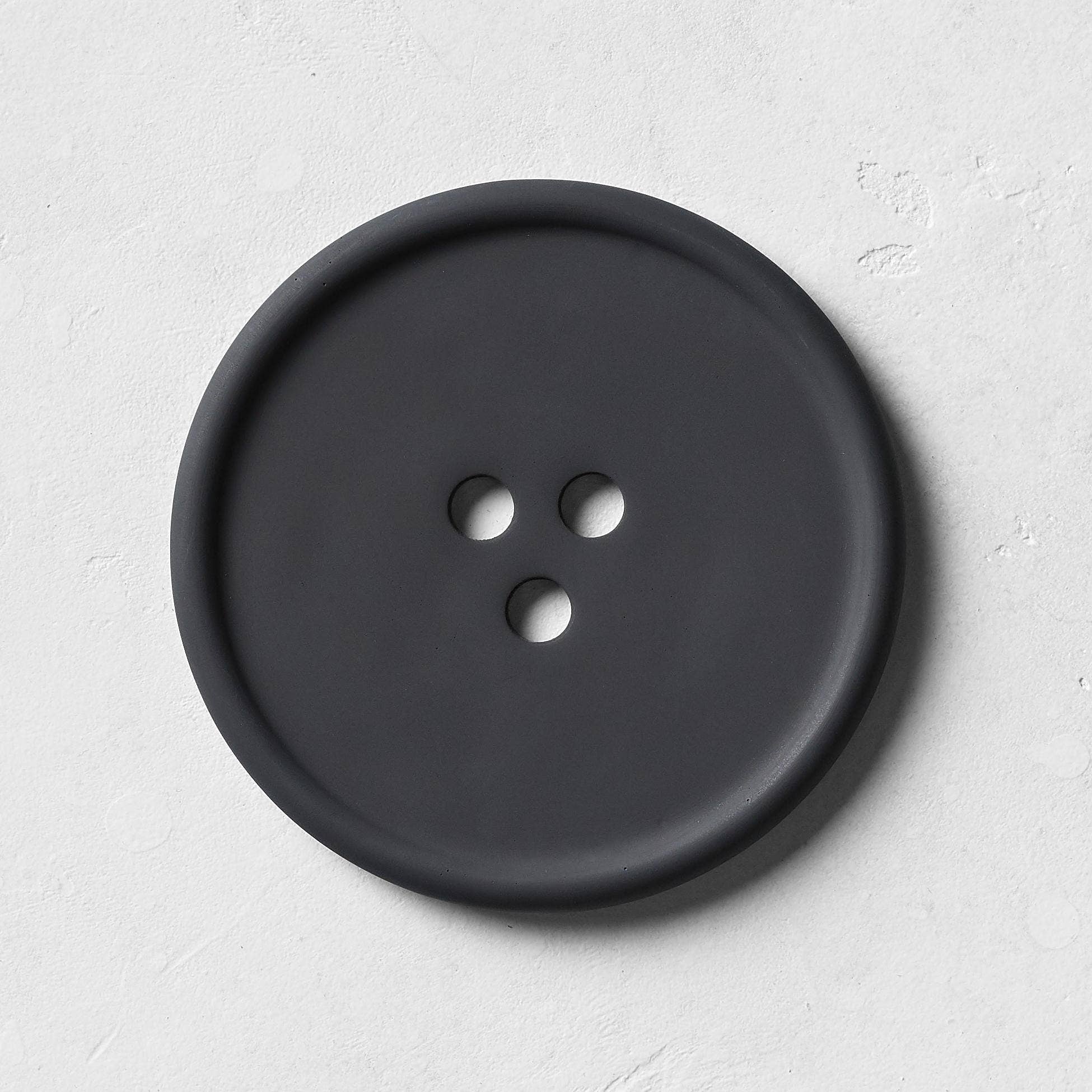 A round black concrete tray with a button-like shape, featuring subtle textures on its surface, designed for holding everyday favorites.
