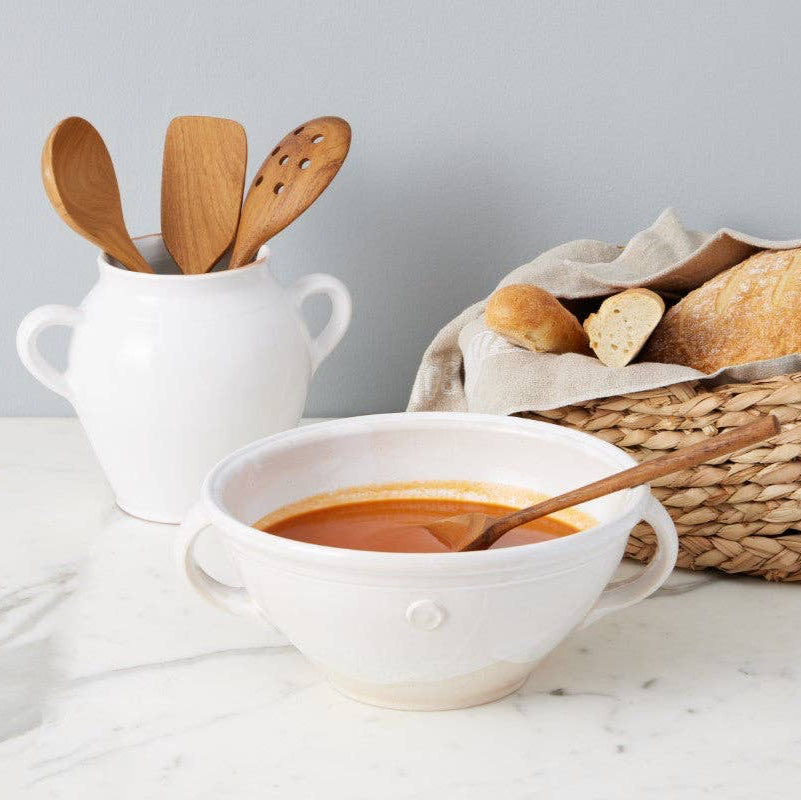 A white mixing bowl full of tomato soup, with wooden spoons and a basket of bread in the background. 