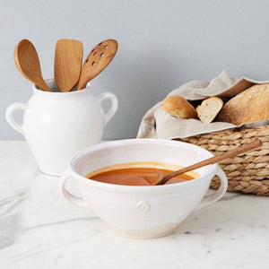 A white mixing bowl full of tomato soup, with wooden spoons and a basket of bread in the background. 