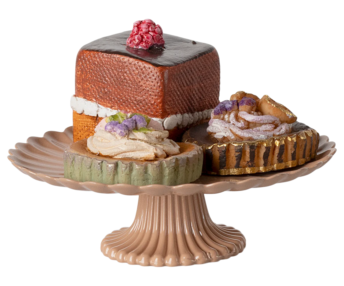 A set of three hand-decorated mini cakes on a rose-colored cakestand, including a chocolate cake, a cheese cake, and a fruit-topped cake.