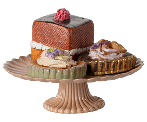 A set of three hand-decorated mini cakes on a rose-colored cakestand, including a chocolate cake, a cheese cake, and a fruit-topped cake.