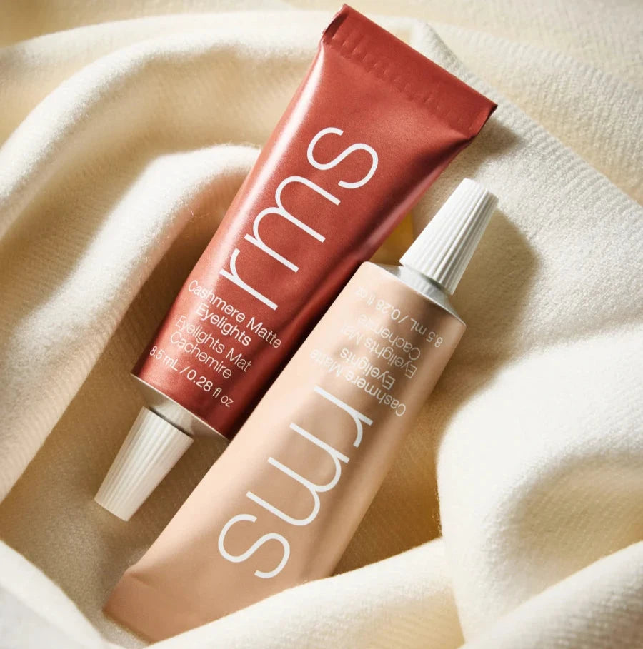Two tubes of RMS Beauty Cashmere Eyelights on a beige fabric surface.