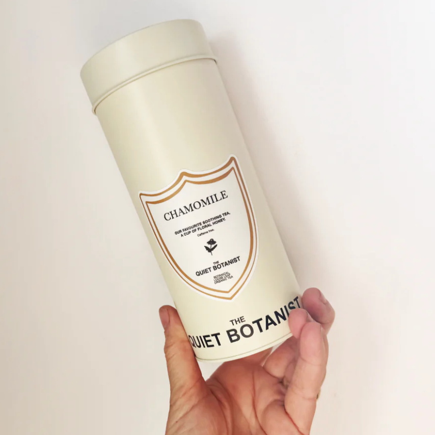 Hand holding a cylindrical container labeled 'Chamomile' by 'The Quiet Botanist' on a light background