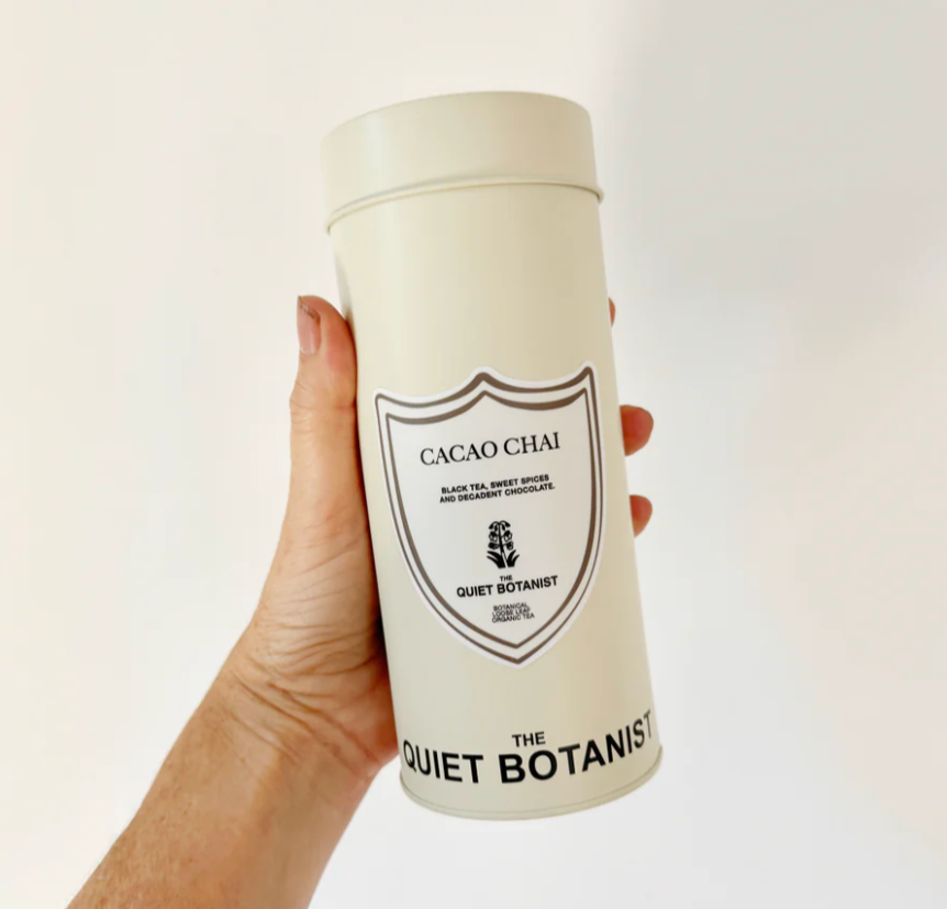 Hand holding a cylindrical container labeled 'Cacao Chai' by 'The Quiet Botanist' on a white background