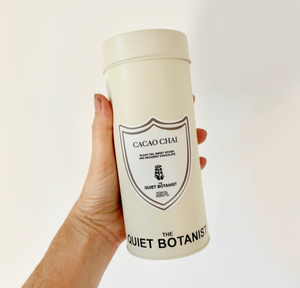 Hand holding a cylindrical container labeled 'Cacao Chai' by 'The Quiet Botanist' on a white background