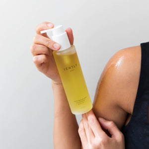 Person applying a yellow bottle of Verto oil to their arm against a light gray background