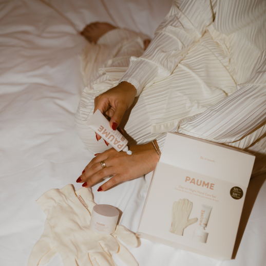 Person holding a bottle of hand cream with gloves and a book titled 'Paume' on a bed.
