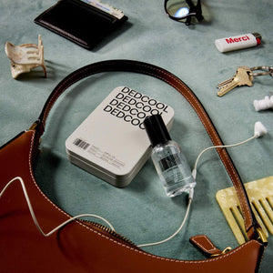 Collection of everyday items including a brown leather bag, bottle, and wallet on a textured surface, with a bottle of Dedcool Taunt and a Dedcool Discovery Set