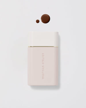 A bottle of Westman Atelier Vital Skincare Complexion Drops with a few drops spilled out on a white surface, showing the product color and consistency.