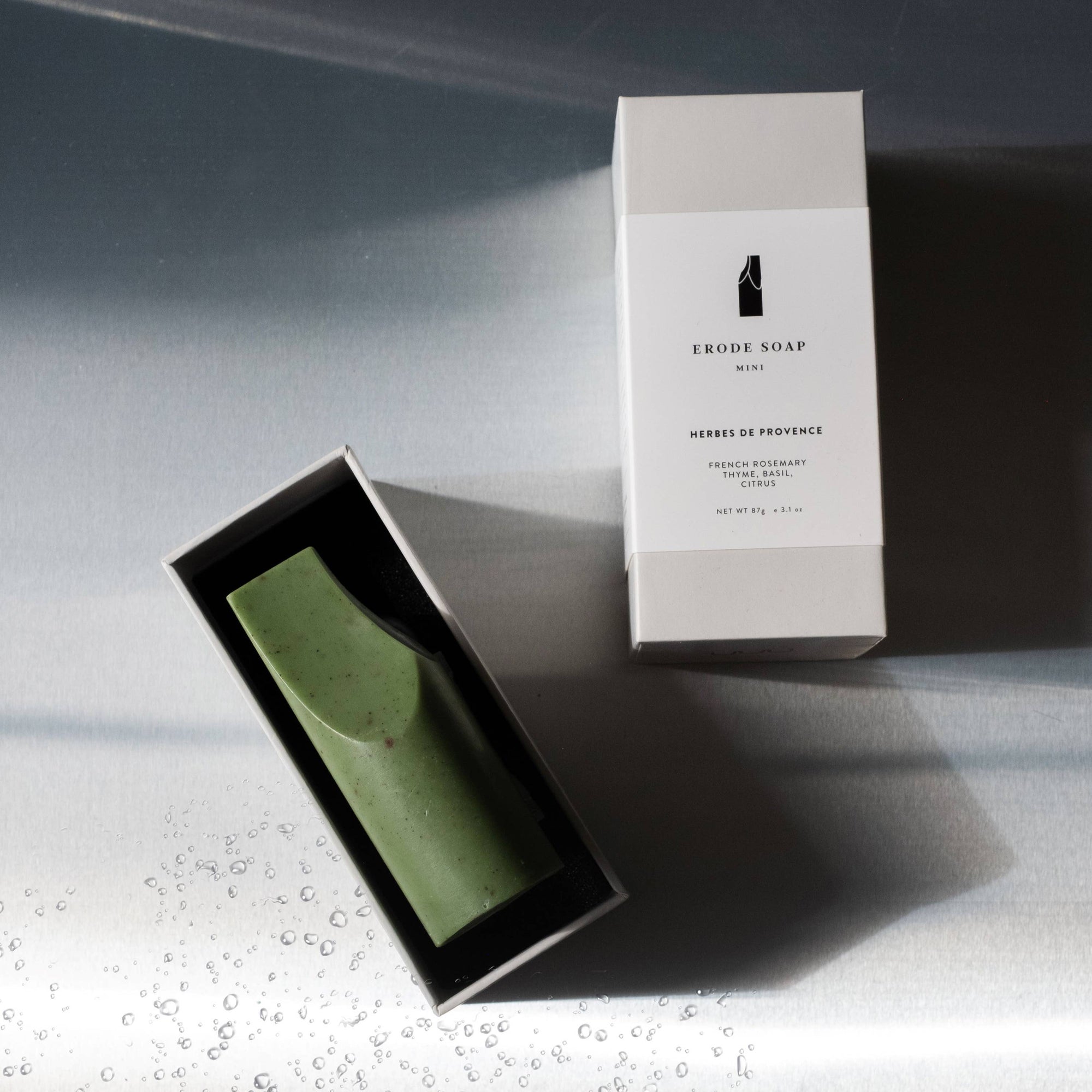 A UMÉ Erode Soap Mini bar next to its packaging box, with a reflective surface in the background.