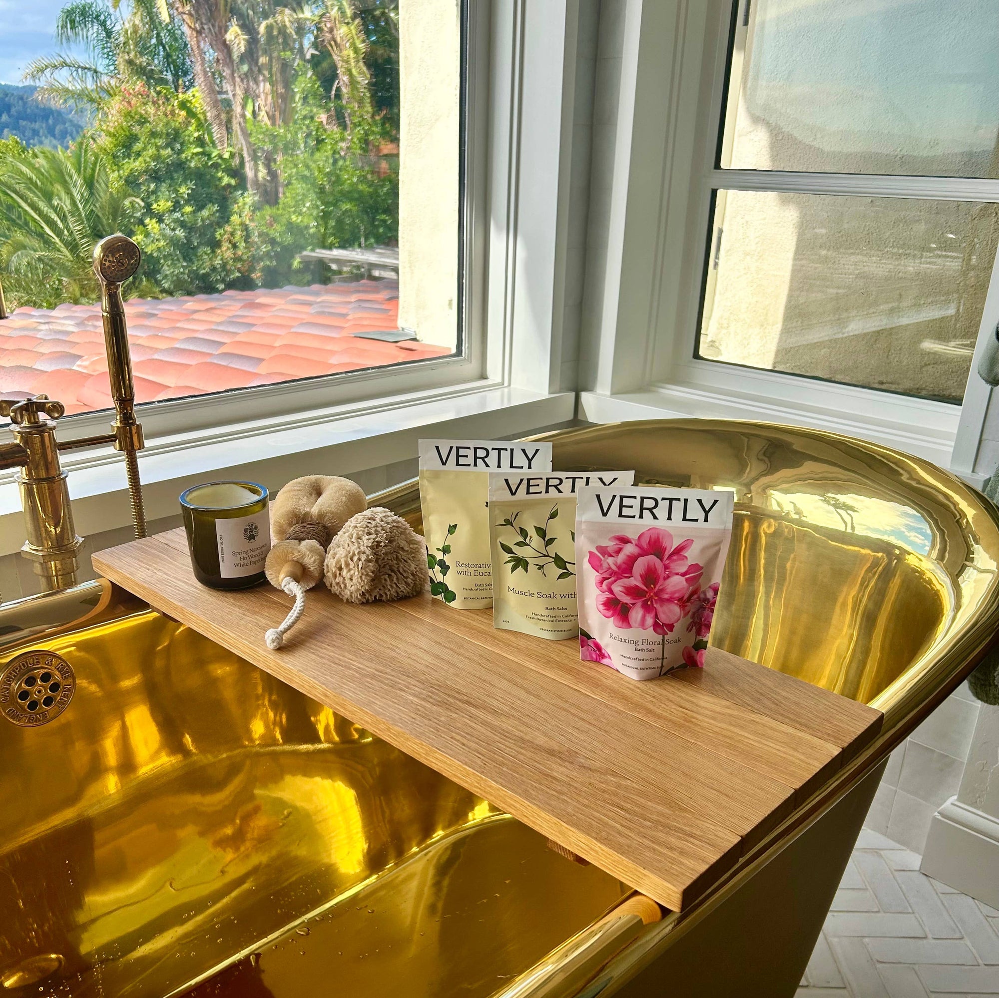 Gold bathtub with a wooden shelf holding bath products near a window with a view of greenery.