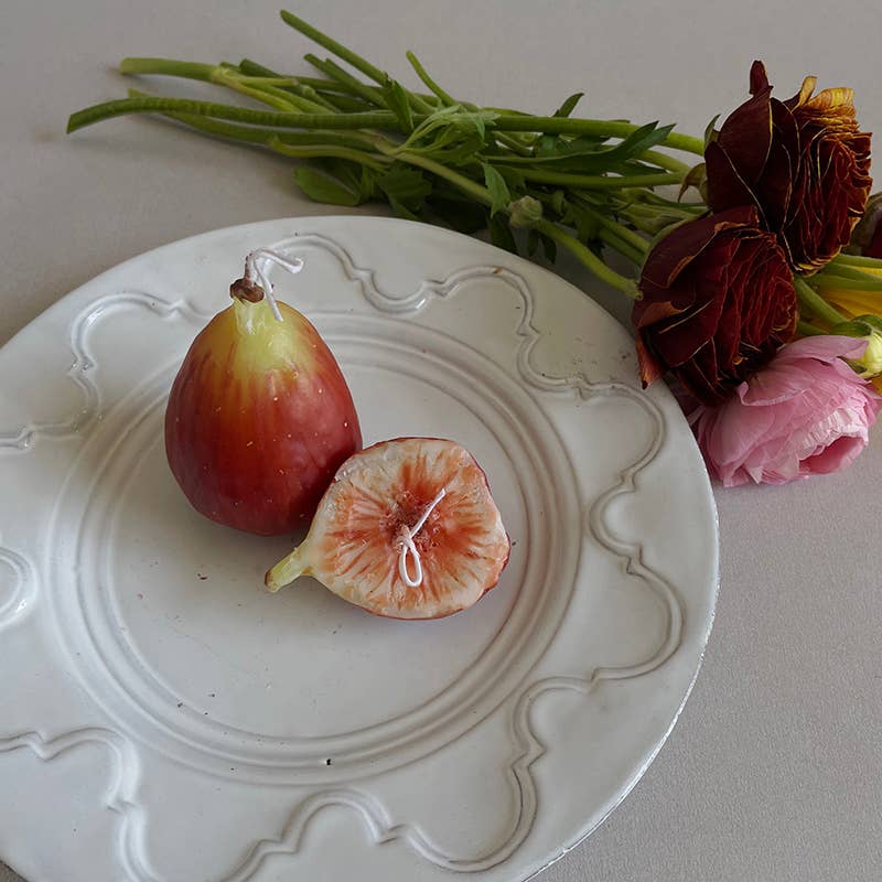 A set of two handcrafted scented candles designed to resemble figs, presented on a white plate with decorative elements in the background.