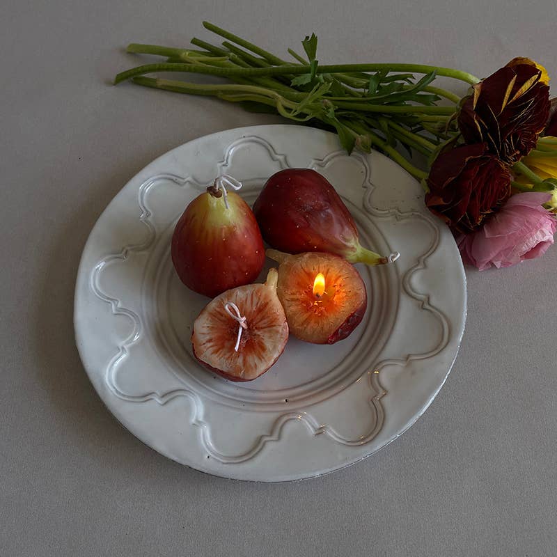 A set of two handcrafted scented candles designed to resemble figs, presented on a white plate with decorative elements in the background.