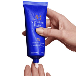 Hand holding a blue tube of Augustinus Bader skincare product on a white background