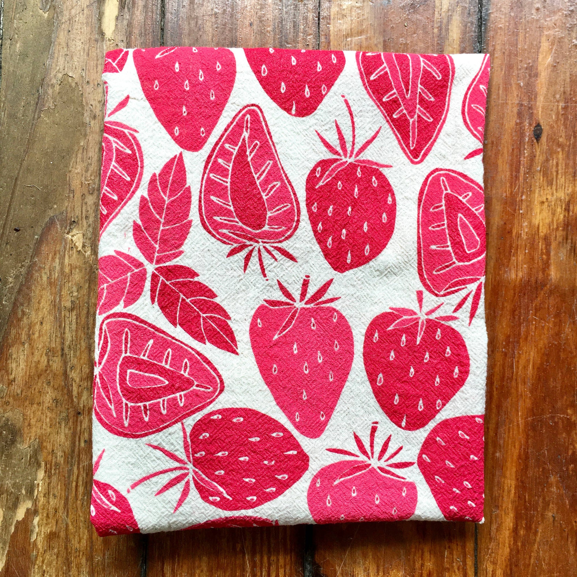 A strawberry block printed tea towel on a wood texture background. 