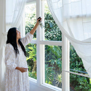 A woman cleaning a large window with white curtains with glass cleaner. 