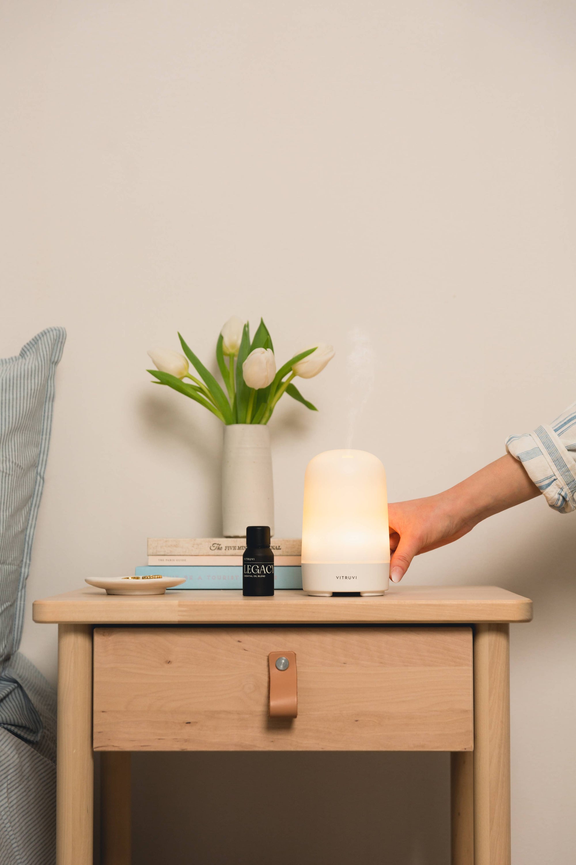 Hand reaching for a Vitruvi Glow diffuser on a wooden nightstand with a vase of flowers and books in the background.