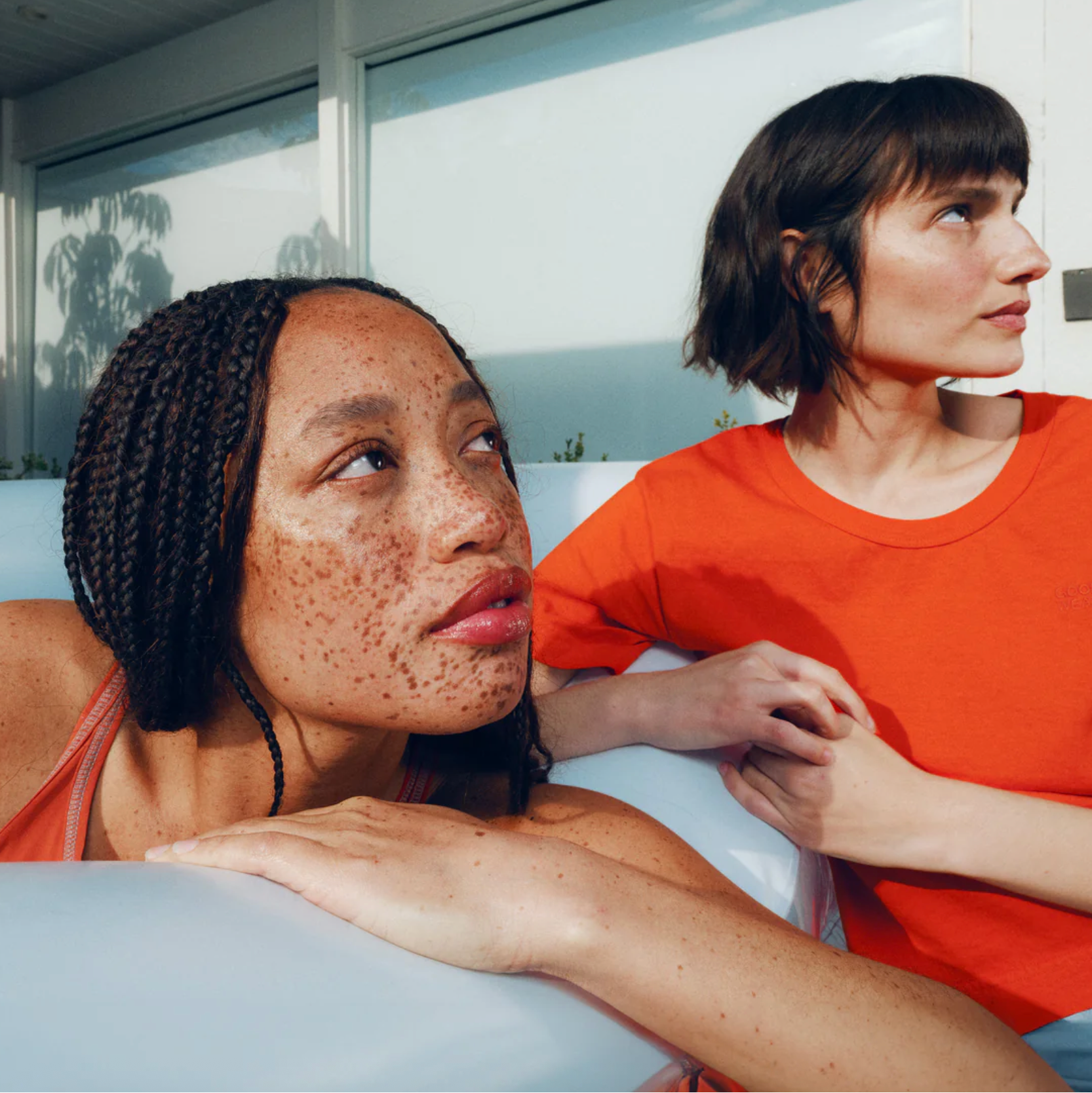 Two women sitting on a couch, one with braided hair and freckles, the other wearing an orange shirt.