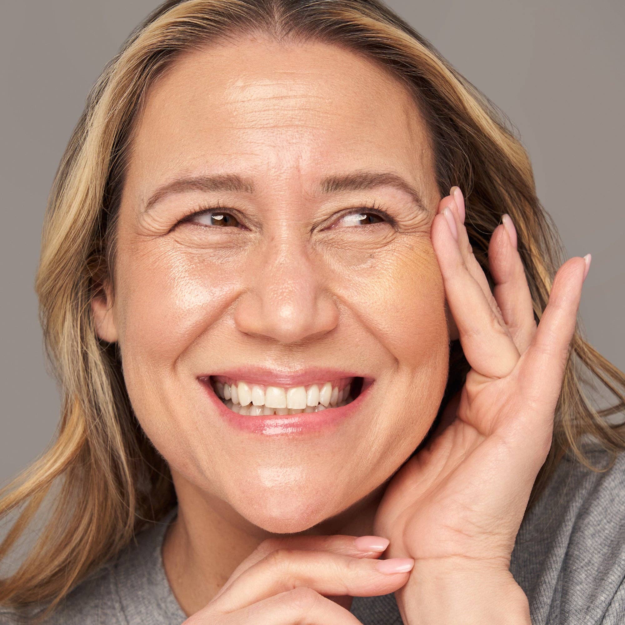 Woman with blonde hair smiling and touching her face against a gray background
