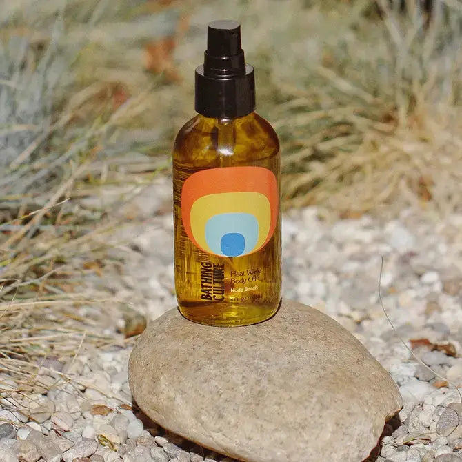 A bottle of Bathing Culture Heat Wave Body Oil with a colorful circular label, placed on a rock outdoors.
