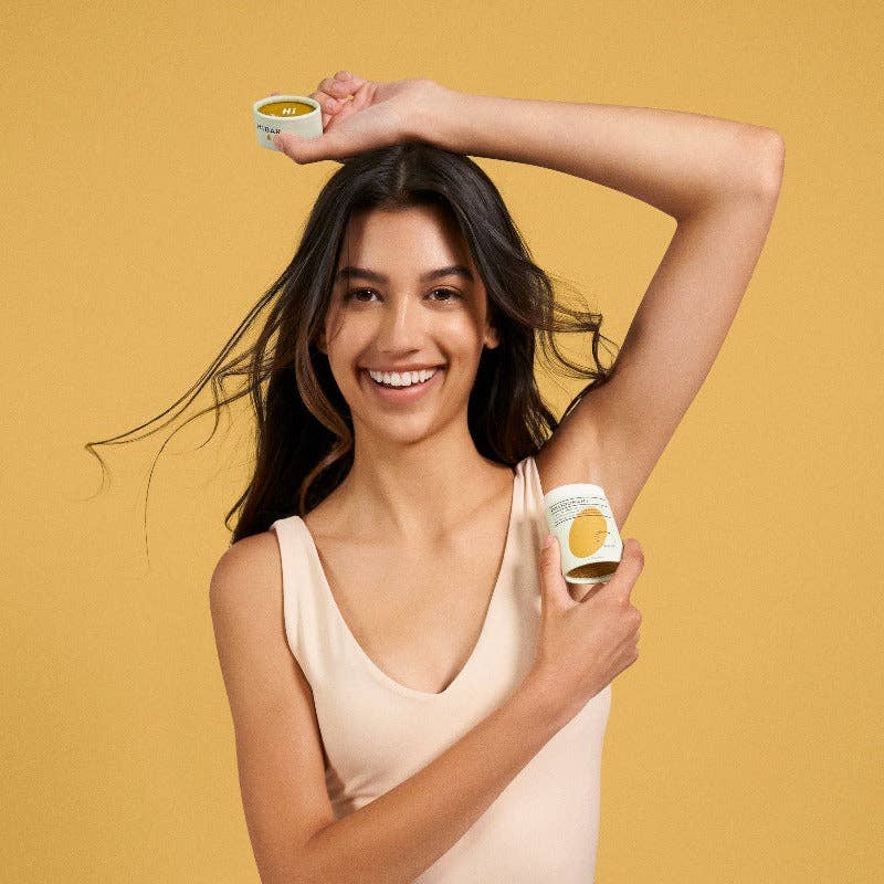 A woman applying a tube of hibar to her underarm on a yellow background. 