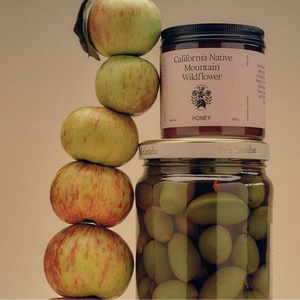 A jar of Flamingo Estate's California native mountain wildflower honey, next to a stack of apples and a jar of olives