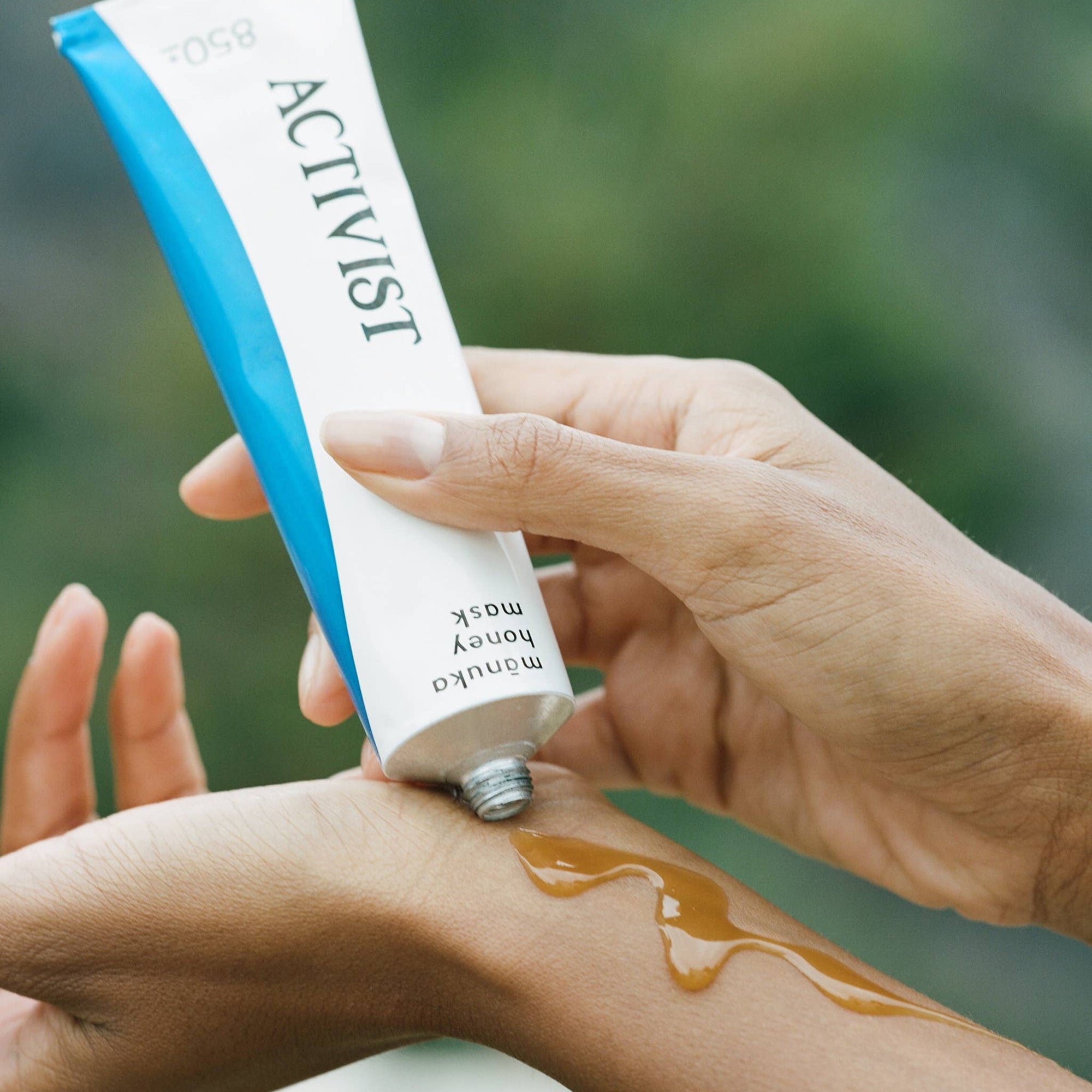 Hand applying honey from a tube labeled 'Activist' to another hand with a blurred green background.