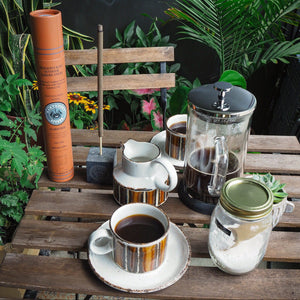 Coffee-making setup with a French press, coffee cup, and other items on a wooden table outdoors.