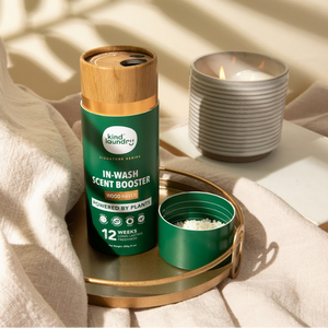 A bottle of Kind Laundry In Wash Scent Boosters placed next to a cylindrical container with a tealight candle on a table with a white linen fabric in the background.