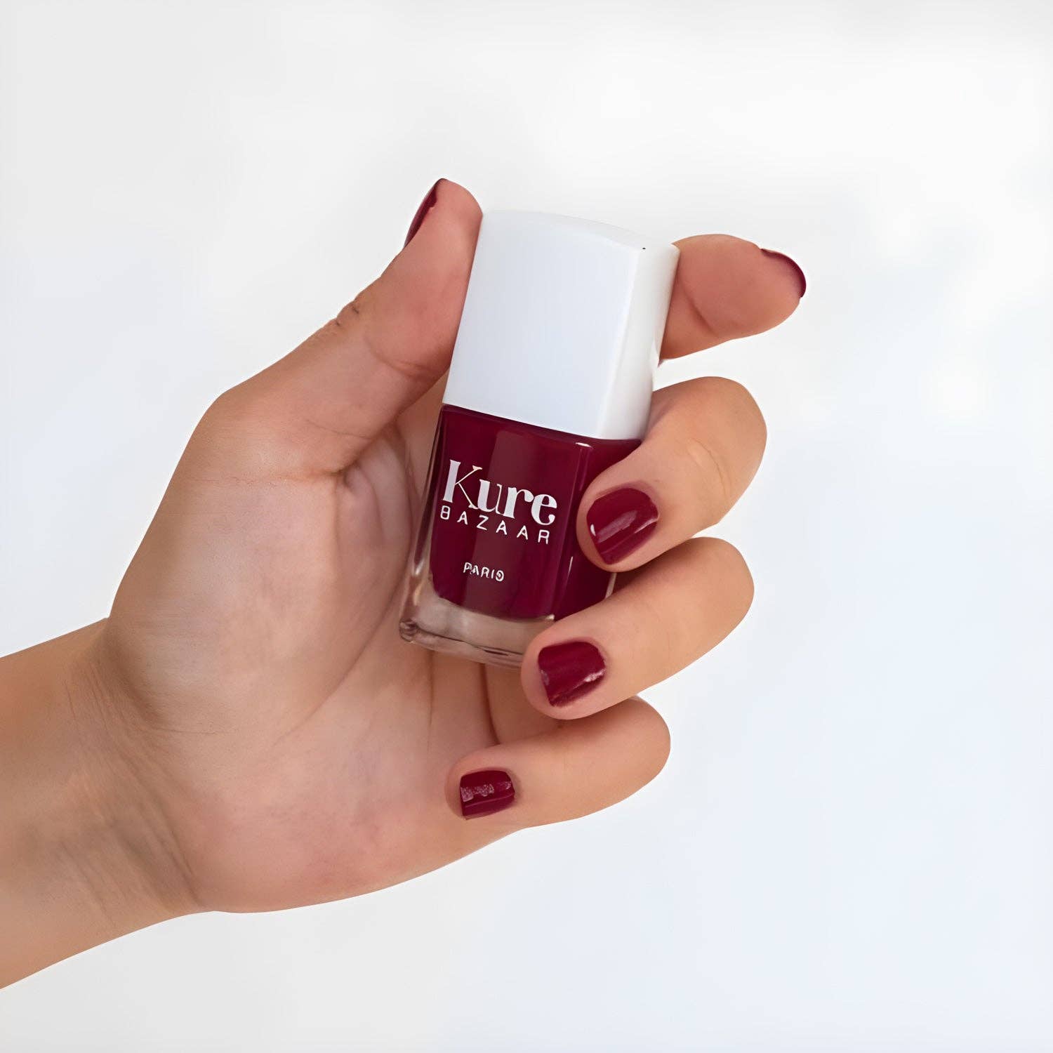 Hand holding a bottle of Kure Bazaar nail polish with a white cap on a white background