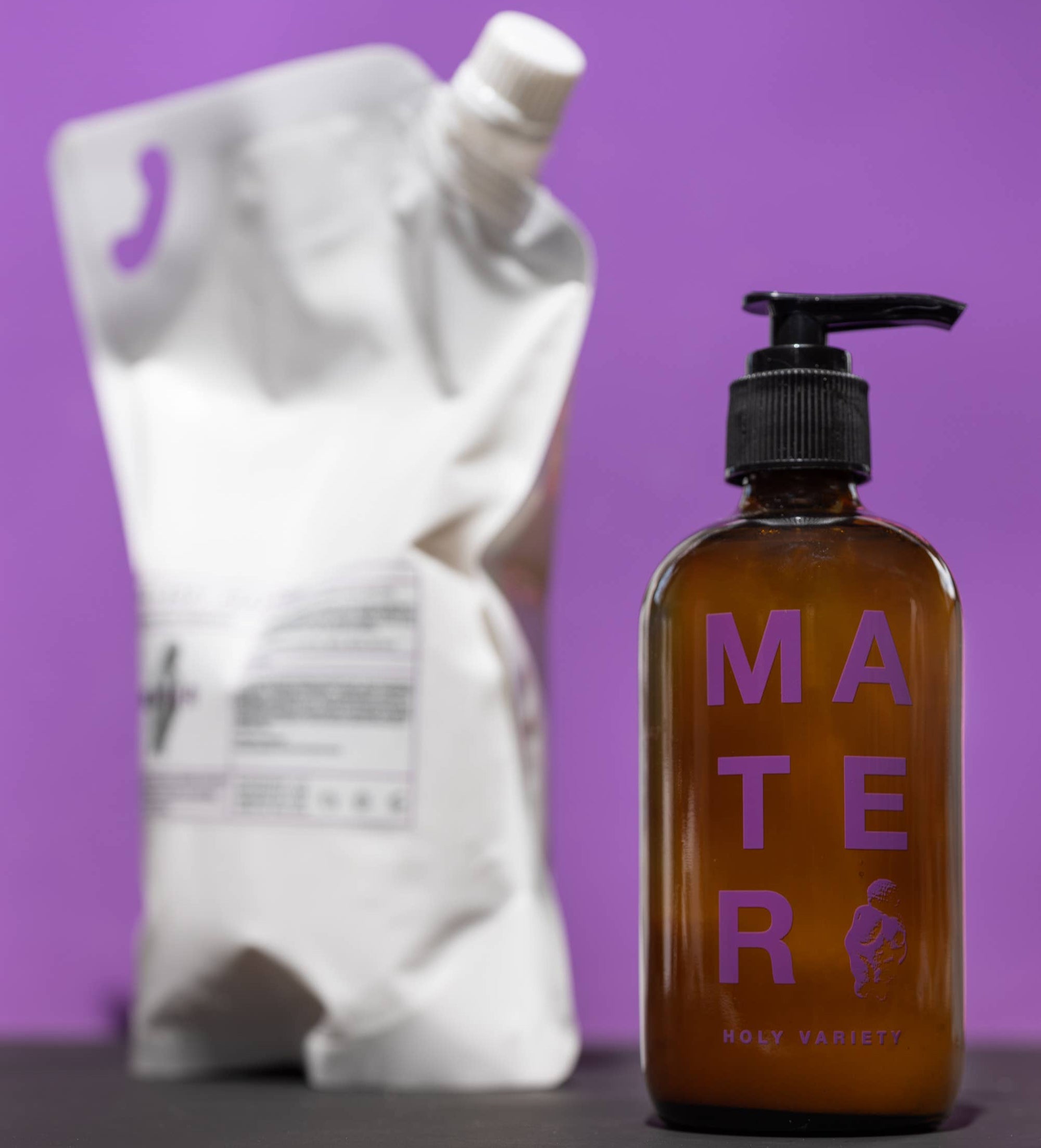 A brown glass bottle with a black pump dispenser labeled 'Mater' with a purple and white text, indicating the 'Holy Variety' line.