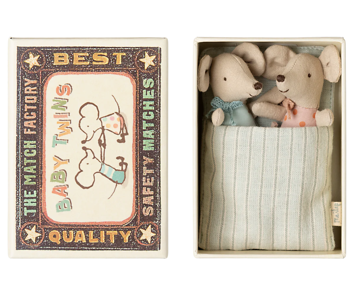 A set of two small toy mice designed to look like baby twins inside an open matchbox. The matchbox is adorned with illustrations and has a striped pattern inside.